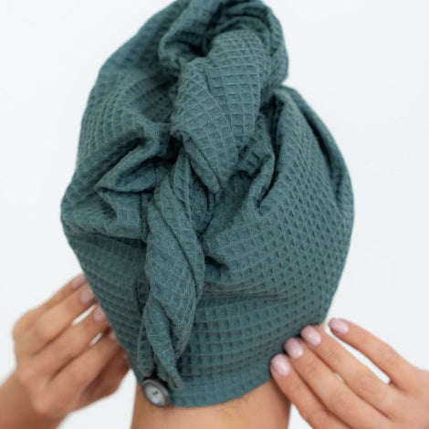 Naisella päässä petroolin sävyinen puuvillainen hiuspyyhe. Pyyhkeessä nappikiinnitys takana.