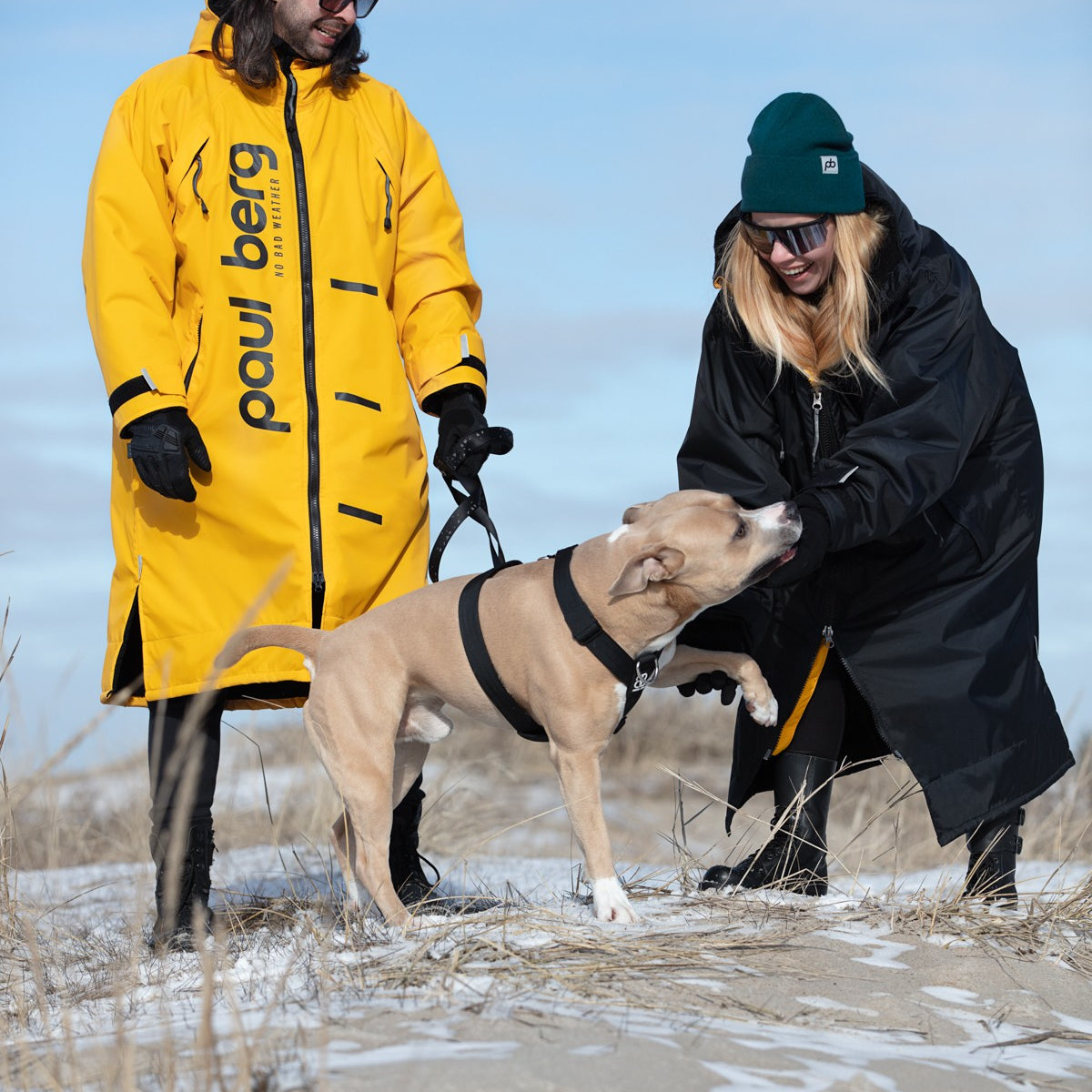 Nainen ja mies ulkoiluttamassa koiraa. Heillä on päällä Paul Bergin monikäyttöiset ulkoilutakit. Miehellä on keltainen ja naisella musta takki.